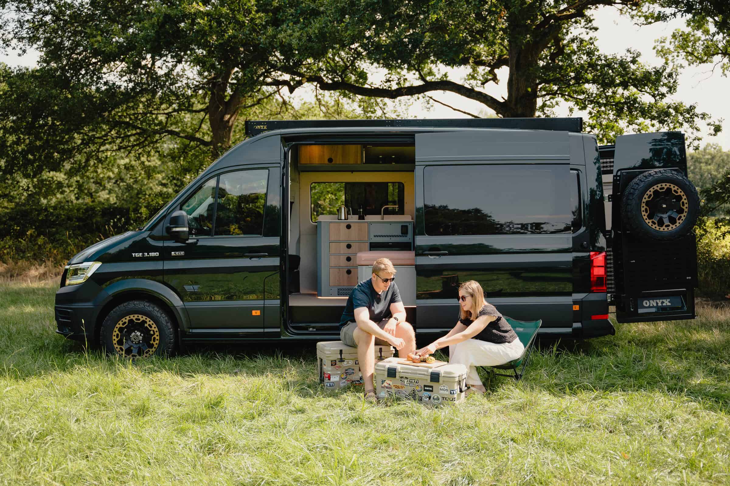 A couple relaxing beside the Onyx Summit campervan in woodland