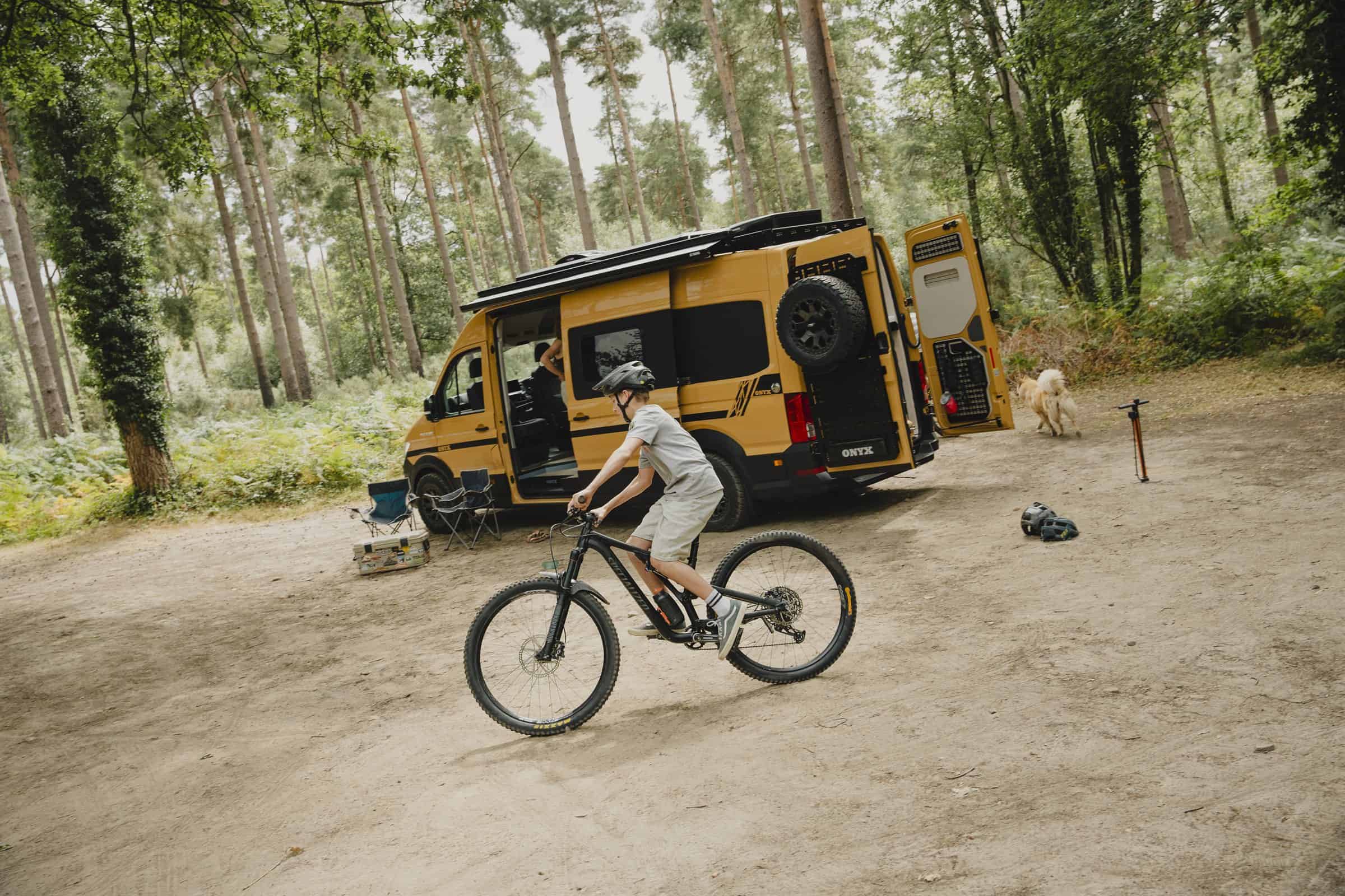 A rider on a mountain bike passing an Onyx campervan parked at a woodland camp