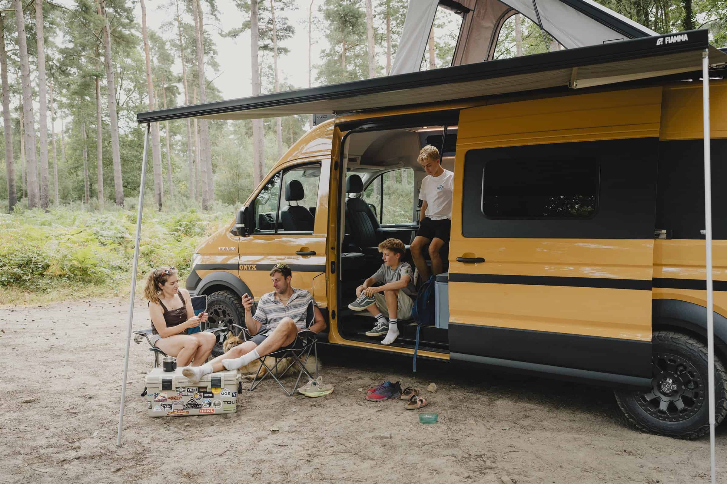 A family of four relaxing under the awning of the Onyx Mesa LWB MAN TGE campervan in woodland, side door open