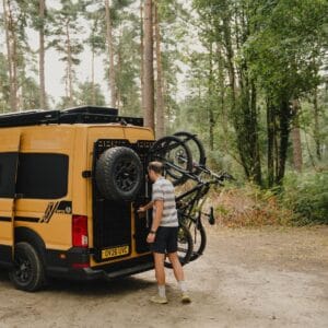 Mountain bikes held upright by 1UP USA racks on the rear door carriers of an Onyx van, beside the spare wheel, in pine woodland