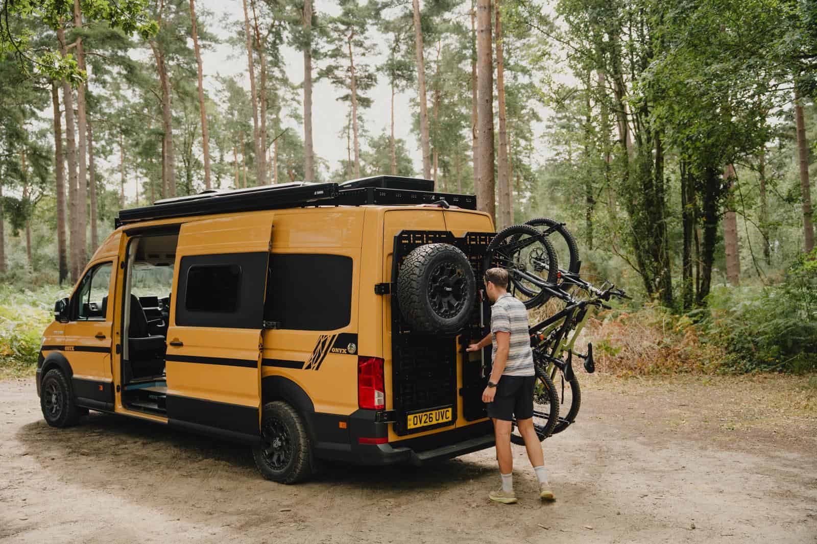 An Onyx van parked in pine woodland with its side door open, while someone loads a bike onto the 1UP USA racks on its rear doors