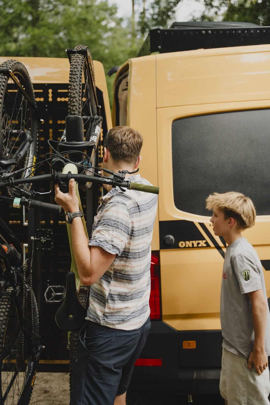 Someone lifting a mountain bike into a 1UP USA rack mounted vertically on the rear door of an Onyx van, while a boy watches