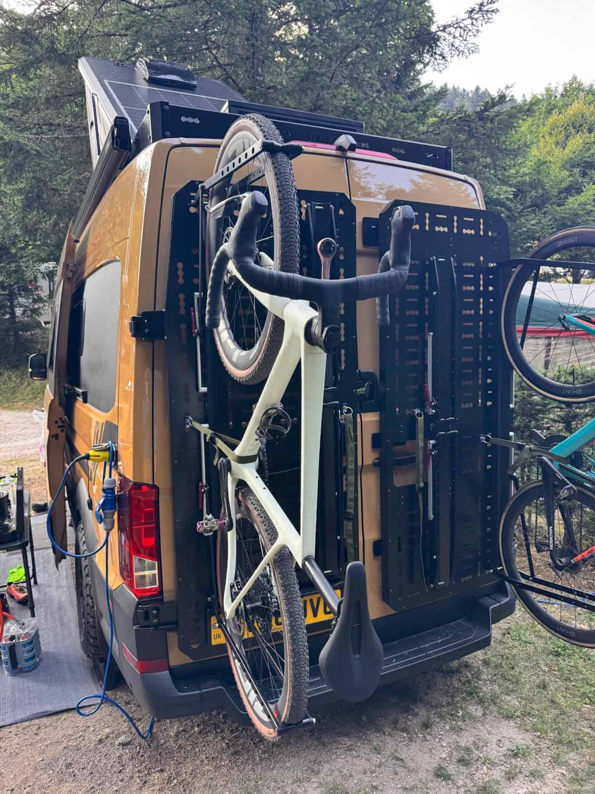 A gravel bike held upright by a 1UP USA rack on the Molle panel of an Onyx van’s rear door, at a campsite