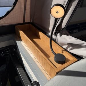 A bamboo storage box mounted inside the Onyx pop-top beside the upper berth, with its reading light switched on