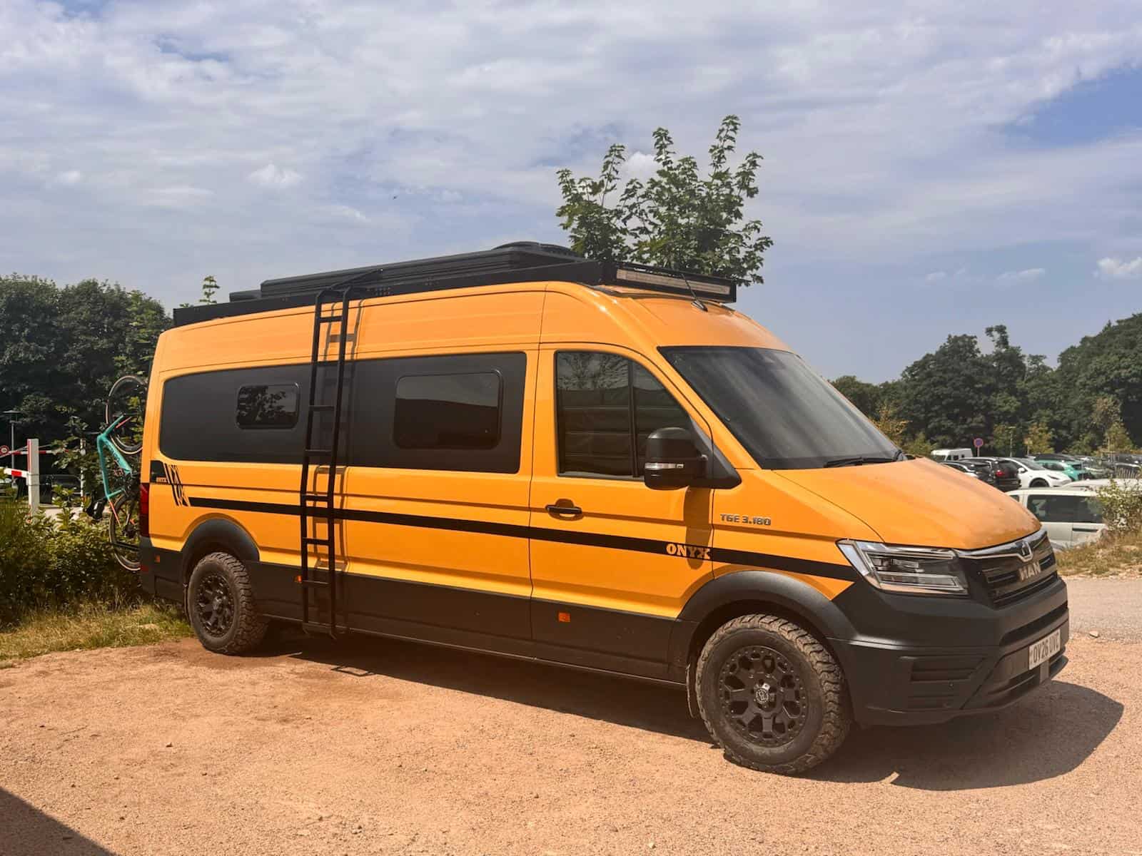 Chris and Louise's amber Onyx van parked on gravel, with roof rack, side ladder and all-terrain tyres
