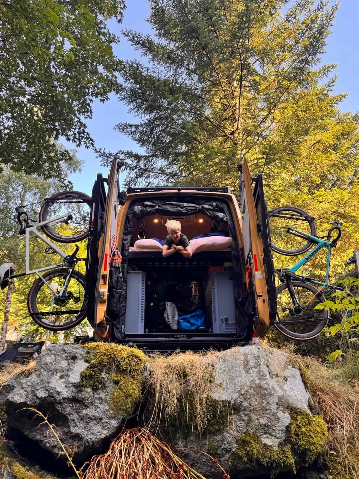 The rear doors of the van open with a mountain bike racked on each one, and a child lying on the bed inside