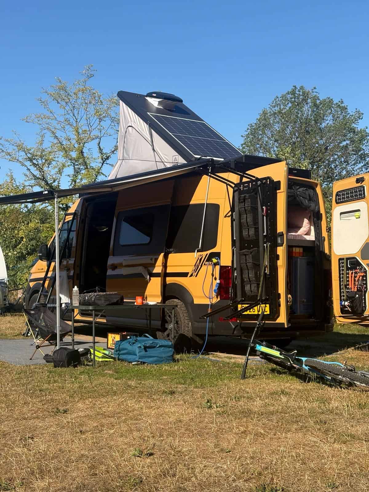 The van pitched at a campsite with the pop-top raised, its solar panel showing, the awning out and kit spread on the grass