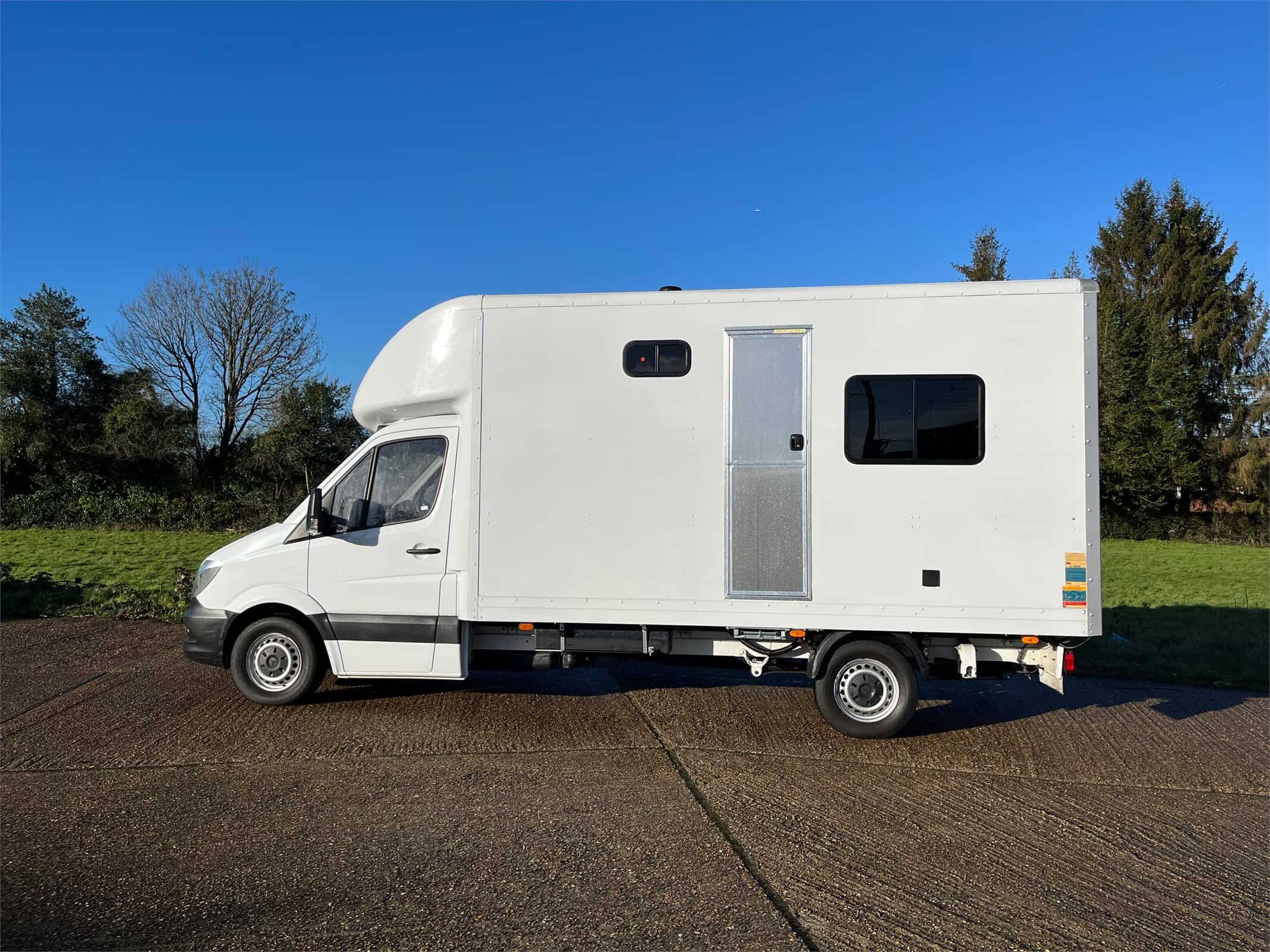 White Luton van with two windows and a door in the body, parked in a green field after its conversion to a campervan.
