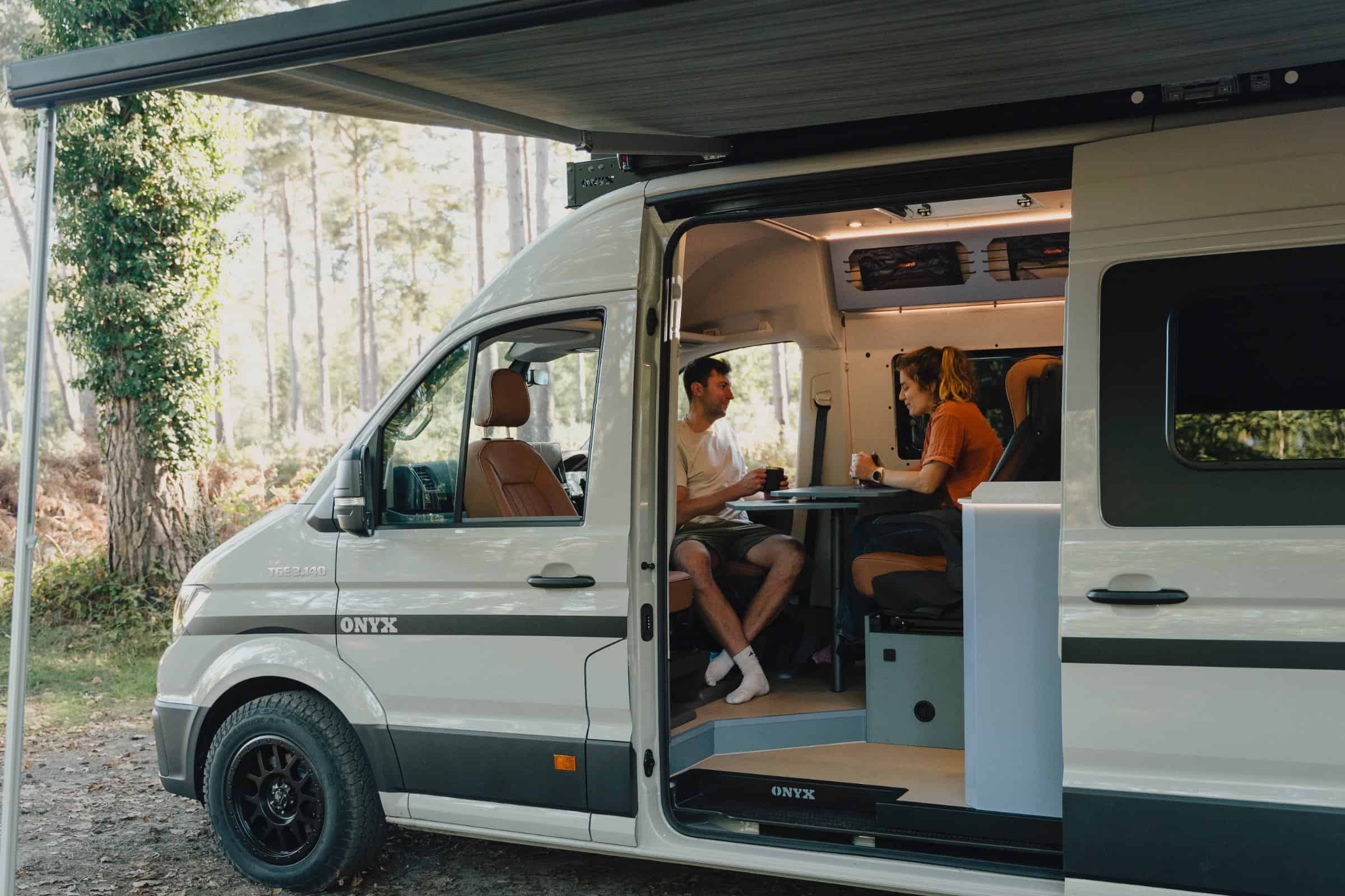 Two people at the table inside the Onyx Mesa campervan, side door open and awning out, in woodland