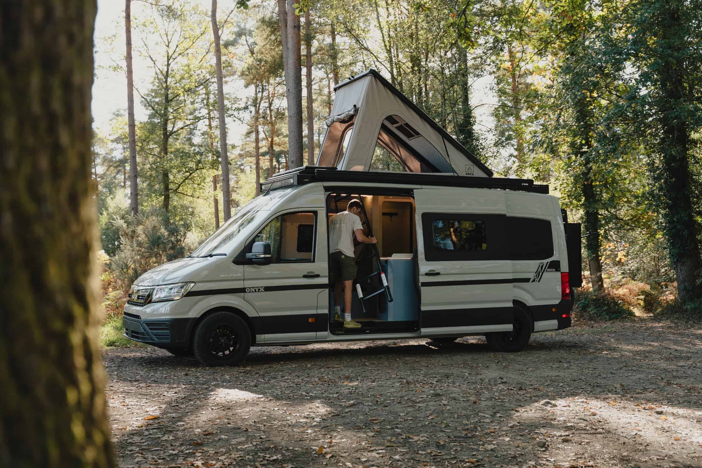 The Onyx Mesa parked among pine trees with the pop-top raised and someone stepping in at the side door